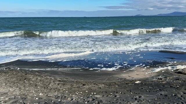 Waves roll onto a black sand beach with foam forming on the shore. The ocean shows shades of blue with a clear sky above. Distant hills are visible on the horizon.