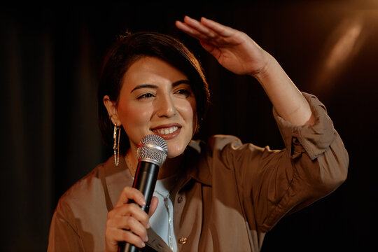 Medium close up of young adult woman performing standup comedy on stage, holding microphone and scanning audience. Useful for entertainment marketing, live event promotion, comedy club ads

