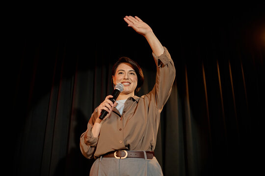 Portrait of biracial middle aged woman performing standup on stage, holding microphone and raising hand. Useful for comedy event promotion, public speaking, entertainment marketing
