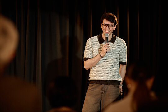Young adult Caucasian man performing standup comedy on stage, speaking into microphone before audience. Useful for entertainment marketing, live event promotion, comedy club ads
