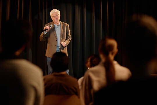 Senior Caucasian man performing standup comedy on stage, speaking into microphone before audience. Useful for entertainment marketing, live event promotion, comedy club ads
