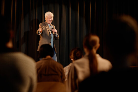 Senior Caucasian man performing standup on stage, speaking into microphone and gesturing toward audience. Useful for entertainment marketing, live event promotion, comedy show advertising
