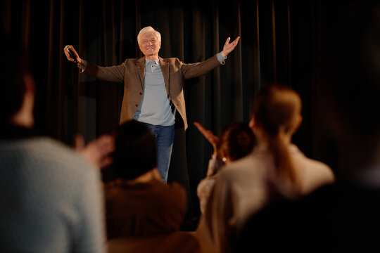 Senior Caucasian man performing standup on stage, greeting audience with microphone and open arms. Useful for comedy event promotion, live entertainment marketing, medium shot
