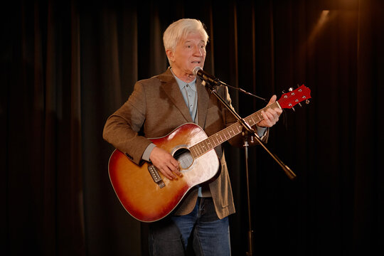 Senior Caucasian man performing standup music set, singing into microphone and playing acoustic guitar on stage. Useful for live entertainment promotion, event marketing, talent ads
