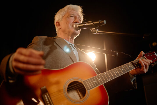 Senior Caucasian man performing standup music set, singing into microphone and playing acoustic guitar on stage. Useful for live entertainment promotion, concert marketing, medium close up

