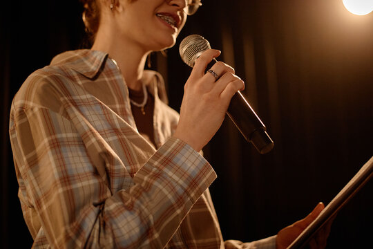 Close up of young adult Caucasian woman performing standup on stage, speaking into microphone while reading notes. Useful for comedy event promotion, live entertainment marketing
