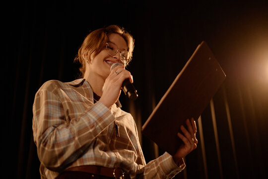 Young adult Caucasian woman performing standup on stage, speaking into microphone while reading notes. Suitable for comedy event promotion, live entertainment marketing, medium close up
