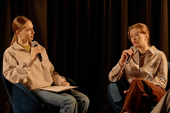 Young adult Caucasian women speaking into microphones during stage interview or standup discussion. Useful for event promotion, public speaking, media training, or live talk marketing
