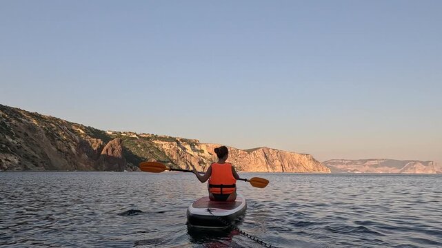 Paddleboard woman sea, person paddling sup board on calm water near a beautiful rocky coast at sunset