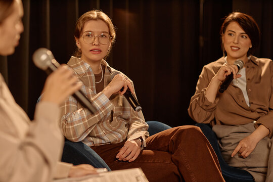 Young adult women speaking into microphones during stage panel discussion, sharing opinions in live standup event. Useful for conference promotion, public speaking, media training

