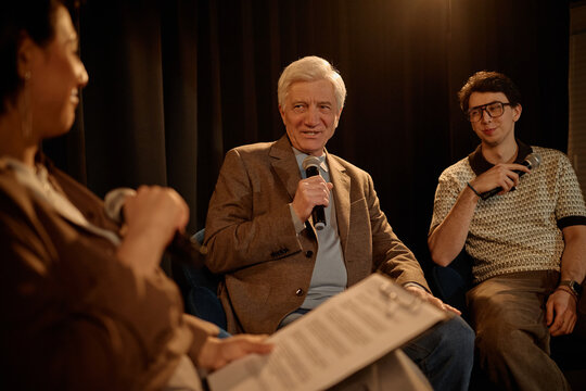 Senior Caucasian man speaking into microphone during stage panel discussion with young adult man, sharing expertise and public speaking concept. Useful for conference, leadership, media event marketin