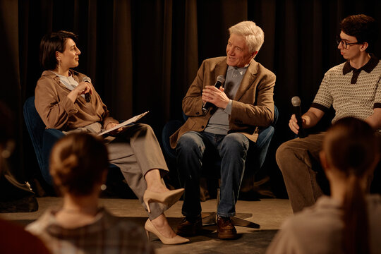 Wide shot of diverse speakers discussing ideas during stage panel interview, holding microphones before audience. Useful for conference marketing, public speaking, live events
