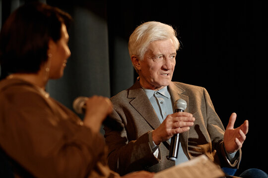 Senior Caucasian man speaking into microphone during stage interview with woman moderator, sharing expertise and leadership insights. Ideal for conference promotion, public speaking, media communicati