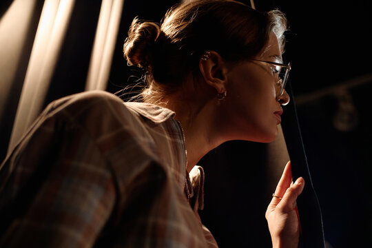Young adult Caucasian woman peeking from stage curtain, preparing for standup performance and checking audience timing. Useful for entertainment marketing, live event promotion
