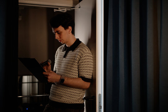 Young adult Caucasian man reviewing notes and holding microphone backstage before standup performance. Useful for comedy event promotion, public speaking, entertainment marketing
