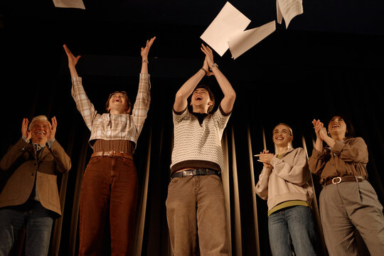 Diverse adults standing on stage, applauding and tossing papers upward after standup performance. Ideal for comedy event promotion, team celebration, public speaking campaigns
