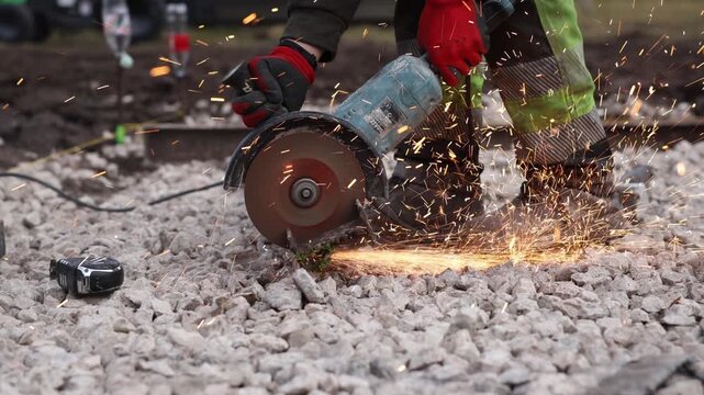 Worker grinds metal rail with angle grinder, sparks arc forward. Gloved hands steady tool over crushed stone near railwork. Title Do not use specific location.
