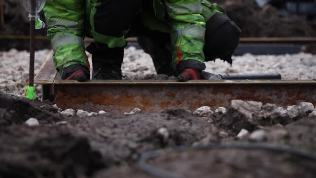 Gloved worker kneels, adjusts taut yellow line on rusted rail, grips hammer, with tape on rebar and bottle marker visible, shallow focus, overcast light.