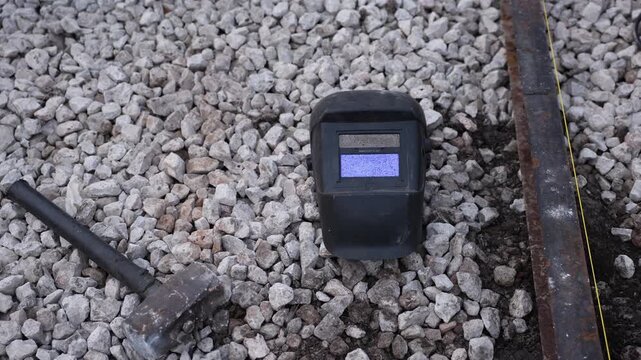 Welding helmet on crushed stone beside sledgehammer and steel rail. Taut yellow string line indicates railwork. Muted metal tones and gravel suggest recent alignment.
