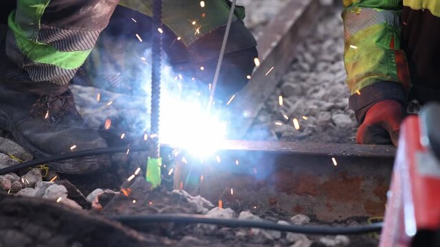 Two workers kneel on crushed stone ballast as one arc welds a rusted rail. White arc, drifting smoke, orange sparks, rebar stake with green tape, daylight, shallow depth.