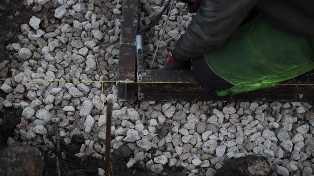 Overhead view shows worker kneeling on crushed stone, using yellow taut strings, a metal clamp, and a tie tool to mark perpendicular lines on a rusted steel frame.