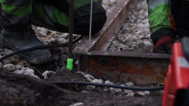 Two workers kneel on gravel ballast and weld a rusted rail as sparks fly, neon green tape marks rebar, red gloves and scuffed boots show on site repair work
