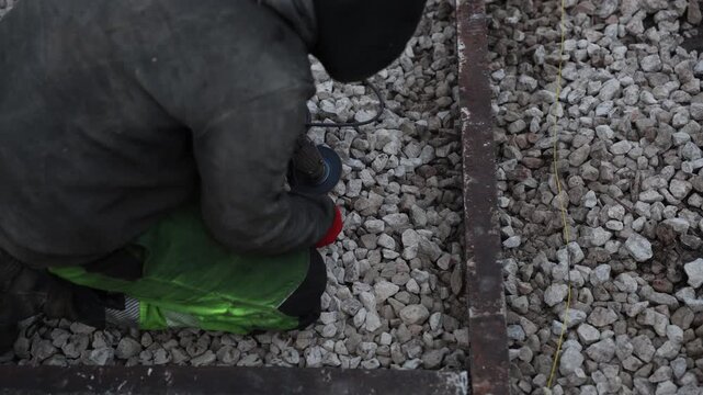 Kneeling worker uses angle grinder on rusty beam set in crushed stone. Yellow guideline runs parallel. Red gloves and green knee pads stand out in muted winter light.