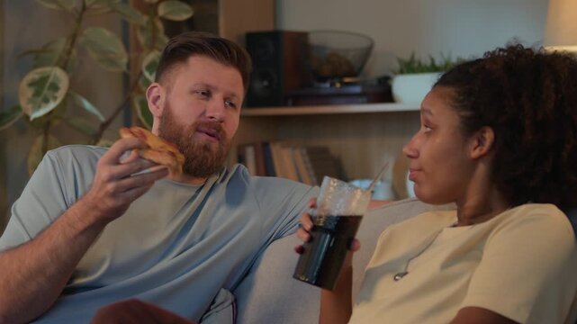 Medium shot of young Caucasian man and Black woman relaxing on sofa in modern living room and chatting while enjoying pizza and drinking soda