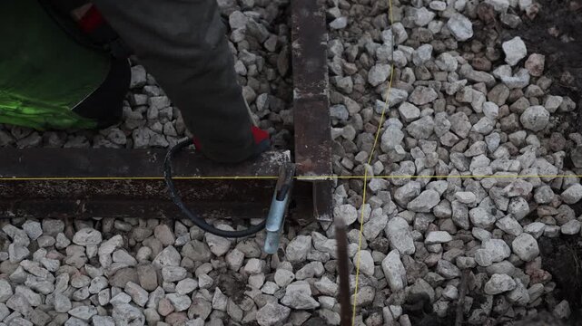 Overhead view shows worker aligning rusted H steel beams on crushed stone as yellow string lines mark layout. Red gloves hold clamp and electrode, sparks show tacking.