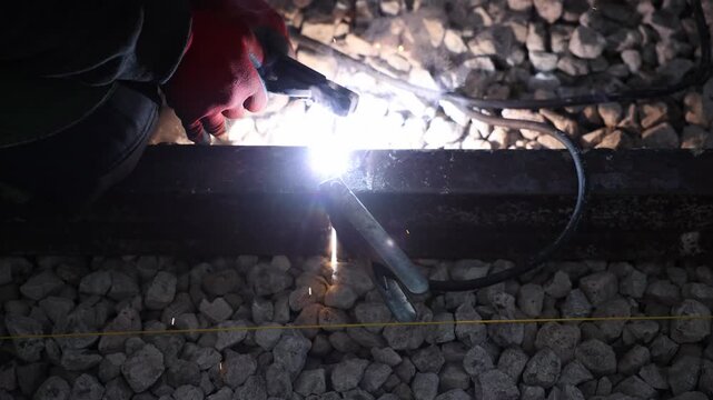 Close up shows a welder's gloved hand guiding an electrode on a steel rail, with bright arc, molten bead, and sparks, heavy grounding clamp, cable, and crushed stone ballast.