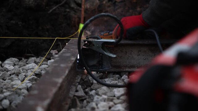 A gloved hand operates a grinder on a rusted steel rail as sparks fly. A clamp and heavy cable rest on crushed ballast. Yellow guide line and weld bead are visible.