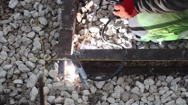 Kneeling worker performs stick welding at a frame joint on crushed stone, with bright arc, sparks, yellow chalk grid, clamp and cable, red gloves, green trousers visible.