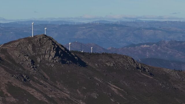 Aerial view of stark white wind turbines contrasting against the rugged, shadowed mountain ridges under a clear sky, Amarante, Portugal.