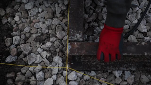 Rusted square steel tubing sits on crushed stone as a red gloved hand adjusts it near intersecting yellow strings, with brief grinder contact during low light exterior work.