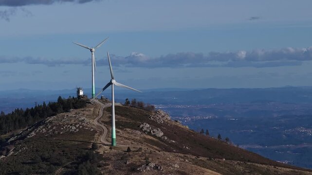 Aerial view of two wind turbines atop a rugged mountain, contrasting with the distant city under a partly cloudy sky, Amarante, Portugal.