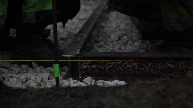 Two workers kneel on crushed stone ballast, align a rusted I beam rail with a taut yellow guide string, sparks flicker as a thin rod suggests welding or grinding.