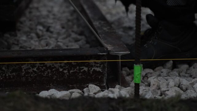 Worker in heavy boots welds rail end at construction site, with smoke and orange sparks, taut yellow string line, and rebar stake with green tape in shallow depth of field.