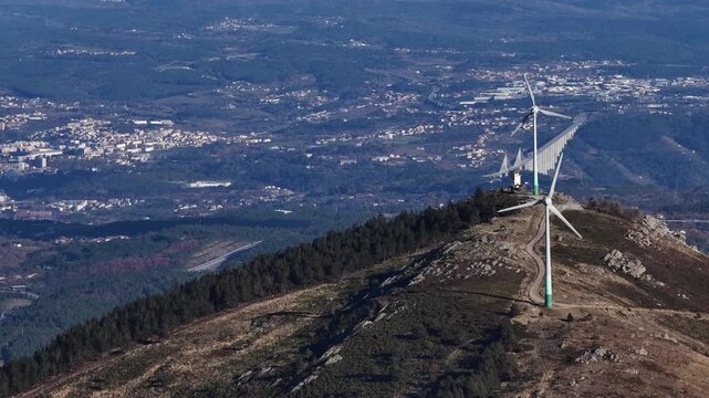 Aerial view of a modern wind turbine atop a mountain with a distant bridge, showcasing the contrast in Amarante, Portugal.