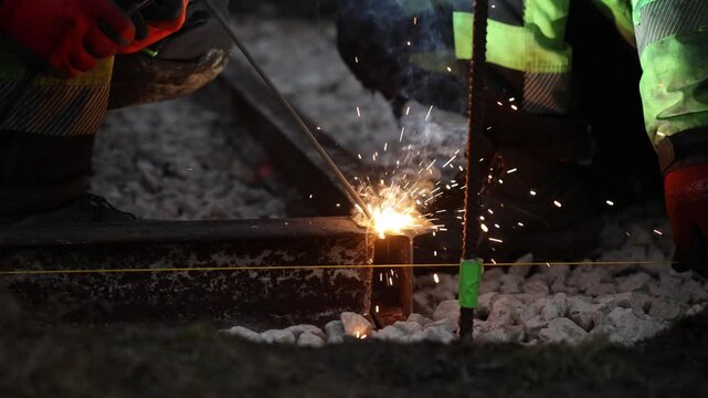 Welders in high visibility jackets fuse a rusted rail at night as arc, sparks, and blue smoke rise over crushed ballast, with green taped rebar and yellow guideline visible.