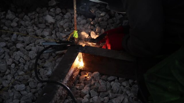 Close up on site welding of steel rail joint on gravel ballast. Red gloves, clamp, rusted rebar with green tape visible as worker manipulates torch and electrode.