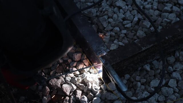 Close welding on rusted rectangular steel frame with sparks and smoke shows steady electrode feed and intermittent sparks, helmet edge, clamp, cables, handheld, shallow depth