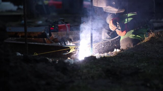 Welder kneels at trench edge, fuses two metal rails with stick electrode, sparks fly, arc light and smoke illuminate gravel, red unit and clamp sit within reach