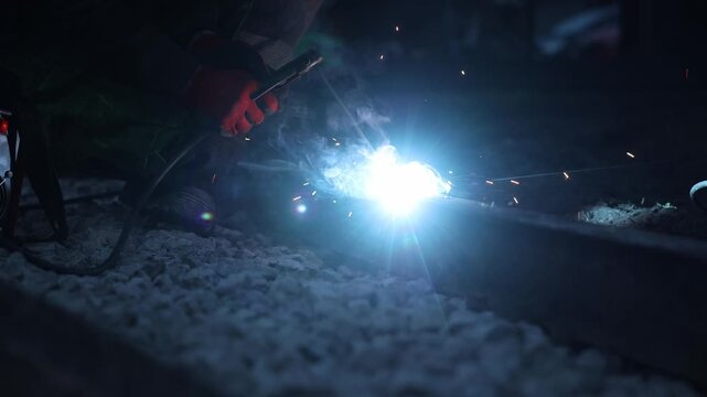 Night shift rail welding shows a worker in red gloves using a torch, steady torch placement and intermittent spark bursts, gravel ballast and cable visible.