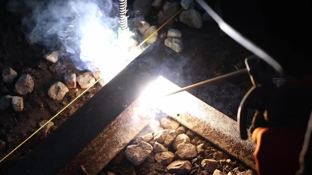 Welder uses stick electrode at night to join rusted steel angles on rocks, with yellow mason line, threaded rebar with green tag, sparks, smoke, and molten droplets.