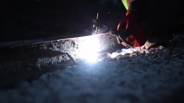 Title  Night rail welding with red gloves and sparks in shallow focus. Technician holds torch to railroad rail, arc throws sparks and smoke over gravel, cool grading, slow shutter.