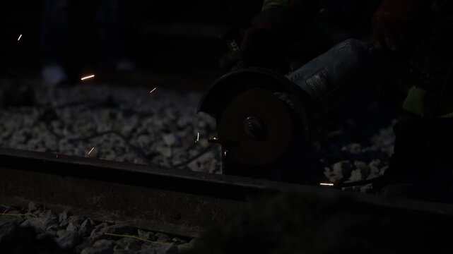 Close low angle view shows rail worker grinding the railhead at night. Bright orange sparks scatter over ballast, boots and power cable near the cutting point.