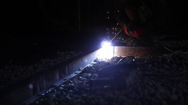 Worker kneels beside rusted rail as arc light flares, sparks jump, and smoke curls. Gloves grip electrode over gravel ballast in low key, shallow depth at night.