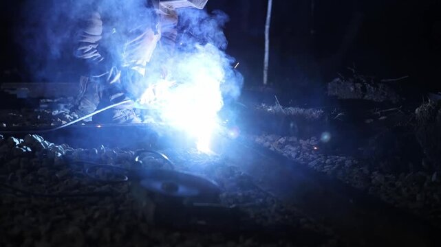 Welder kneels on rail as arc weld sparks and smoke rise at night. Title appears here. Hand moves, molten metal flows, gravel ballast and tools sit near, high contrast cool cast.