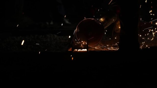 Worker slices steel rail at night with circular disc, sparks trail along track bed. Low key lighting, operator silhouette, visible rail profile and grinding jig.