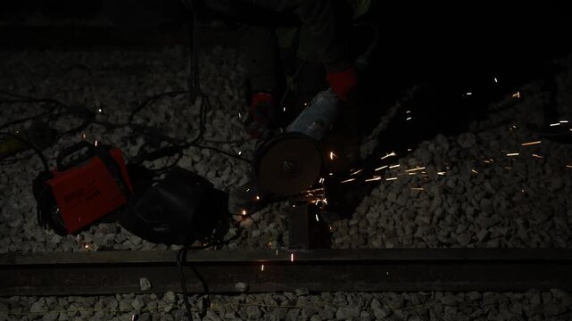Night scene shows railway maintenance worker cutting track with a handheld cutoff saw. Sparks fan across ballast stones. Red gloves and dark welding hood are visible.